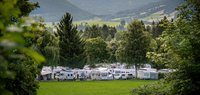 Campingplatz Seewang in Rieden mit Wohnwagen und Zelten auf einer Wiese, umgeben von Bäumen und Bergen im Hintergrund unter bewölktem Himmel.