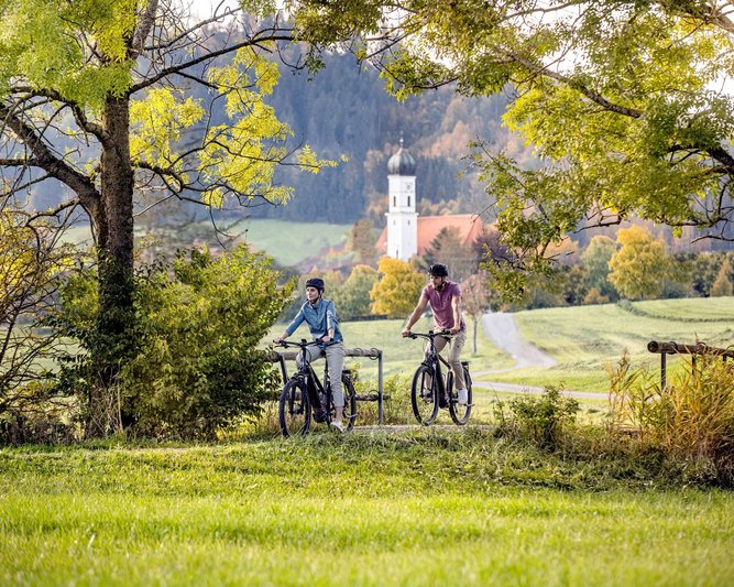 Zwei Radfahrer auf einem Weg im Grünen mit Bäumen im Vordergrund und einer Kirche im Hintergrund in Eisenberg im Allgäu