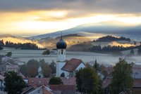 Blick auf Roßhaupten mit Kirche und Zwiebelturm vor nebligen Hügeln im Sonnenaufgang.