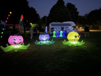 Drei leuchtende, bunte Luftballon-Oktopusse mit großen Augen und lächelnden Gesichtern auf einer Wiese bei Nacht, im Hintergrund Wohnwagen und Menschen.